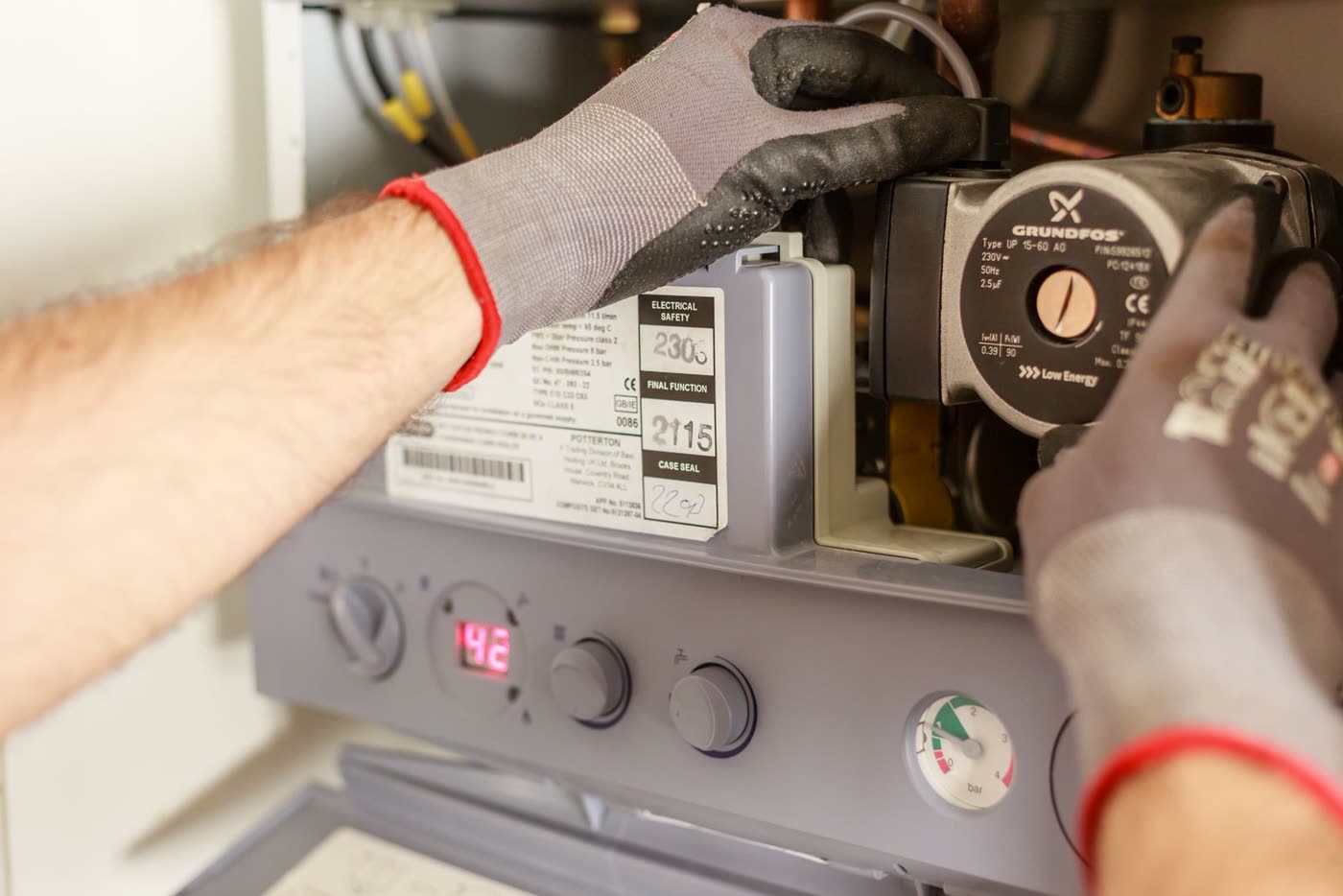 Hände in Arbeitshandschuhen an der offenen Gastherme, Pumpe und Manometer sichtbar