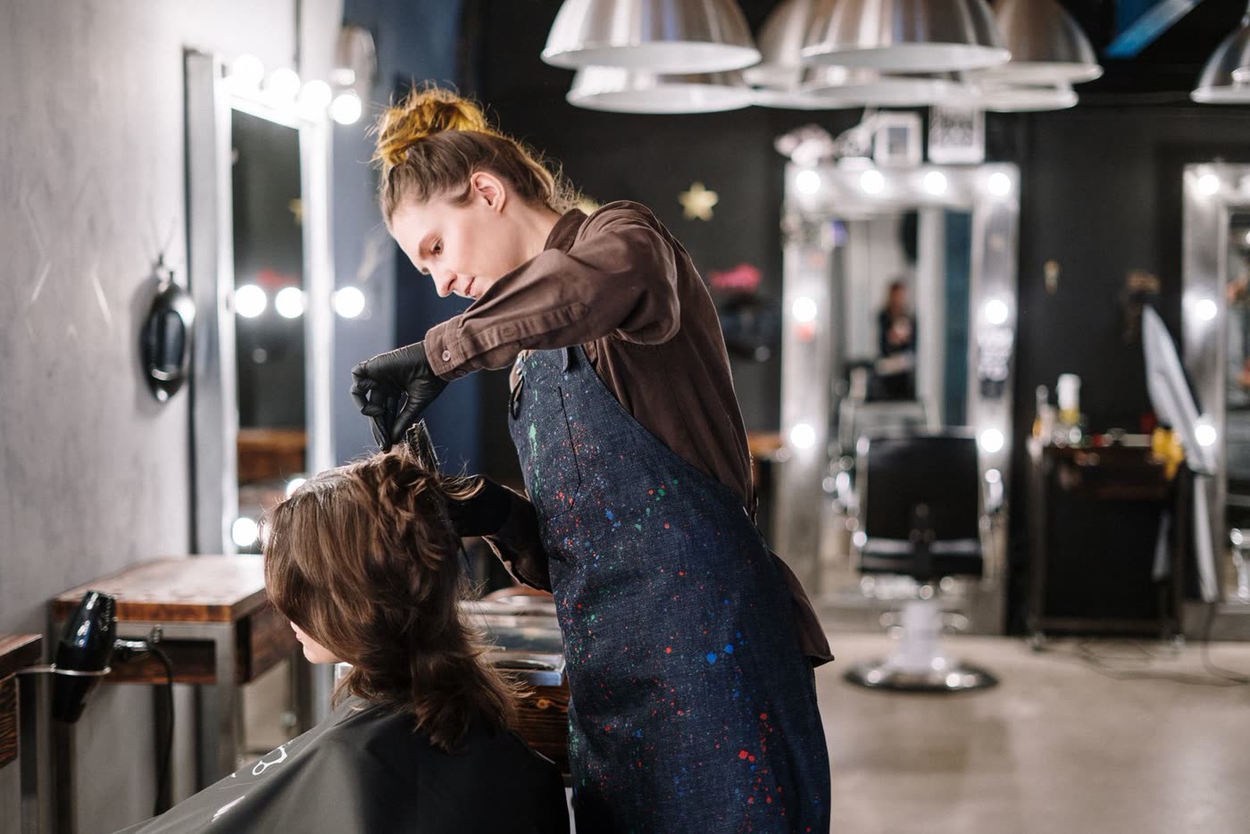 Friseurin in dunkler Schürze arbeitet am Haar einer Kundin, Spiegel mit Glühlampen im Hintergrund